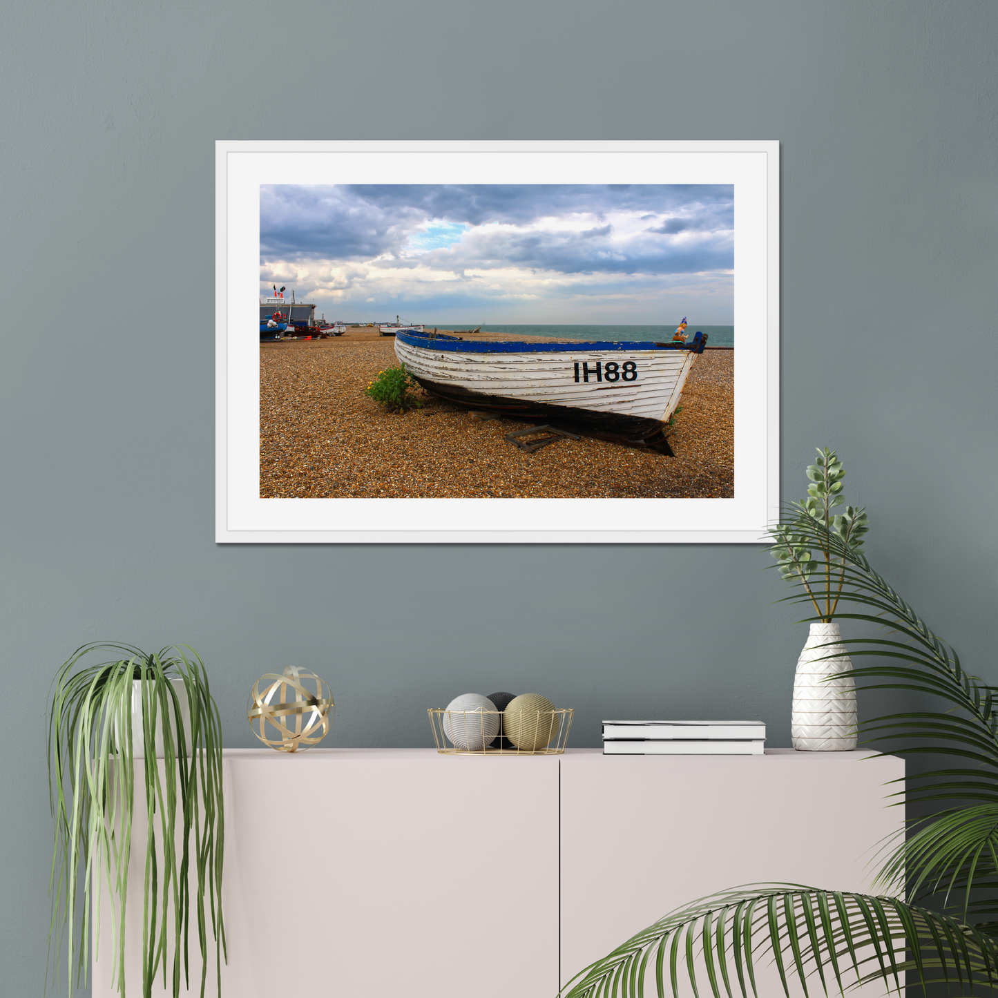 Framed photograph of a boat on a pebbly beach with a cloudy sky on a gray wall above a white cabinet with decorative items.