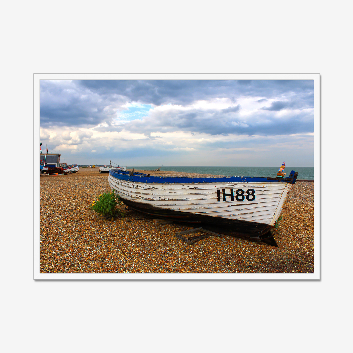 Framed photograph of a boat on a pebbly beach with a cloudy sky.