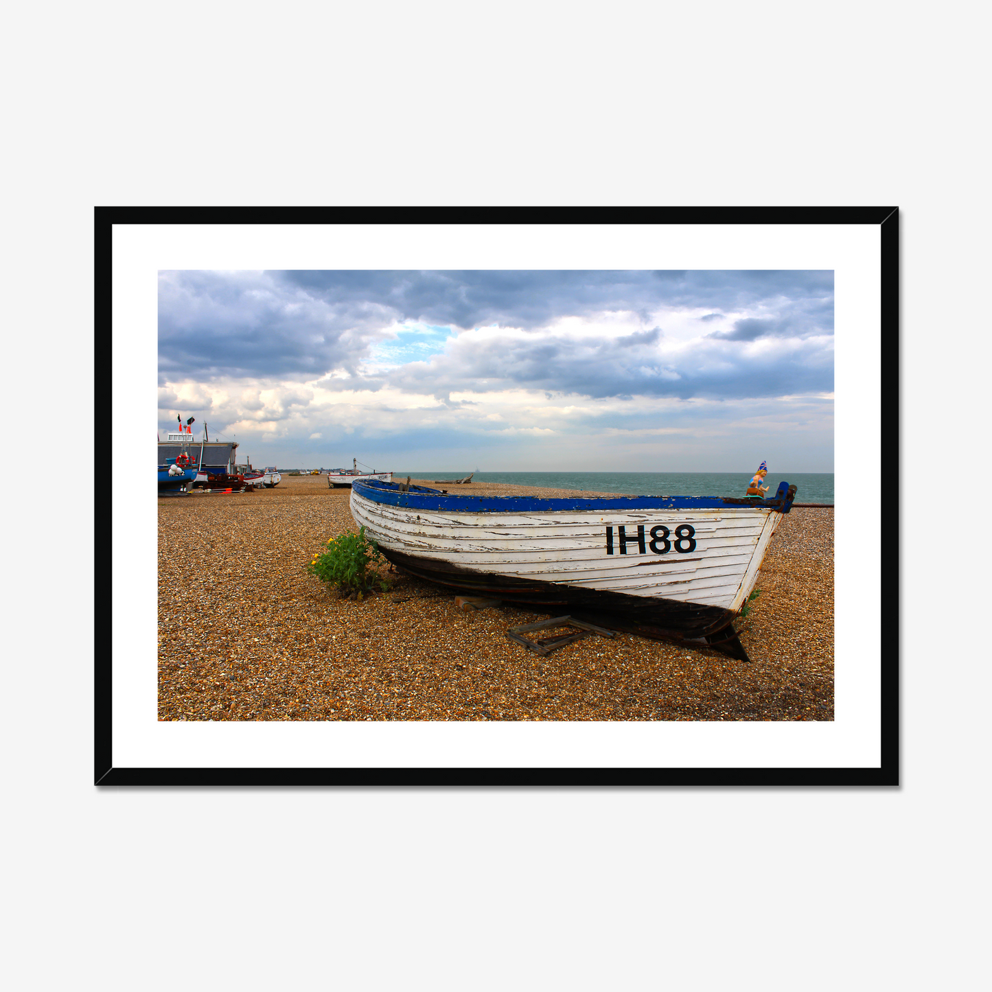 Framed photograph of a boat on a pebbly beach with a cloudy sky.