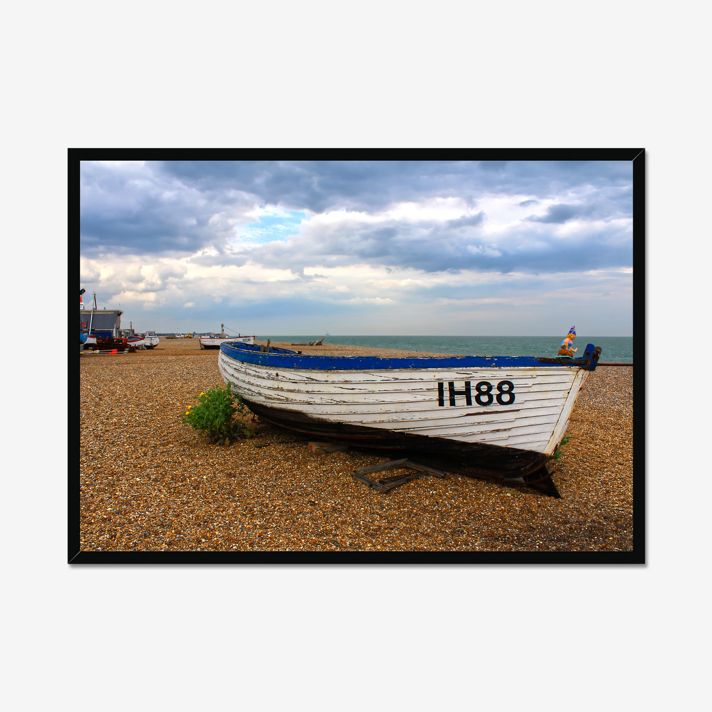 Framed photograph of a boat on a pebbly beach with a cloudy sky.