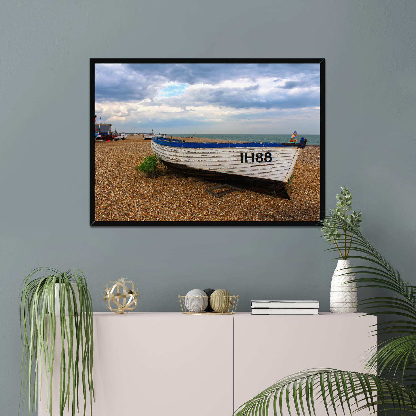 Framed photograph of a boat on a beach hanging on a wall above a white cabinet with decorative items.