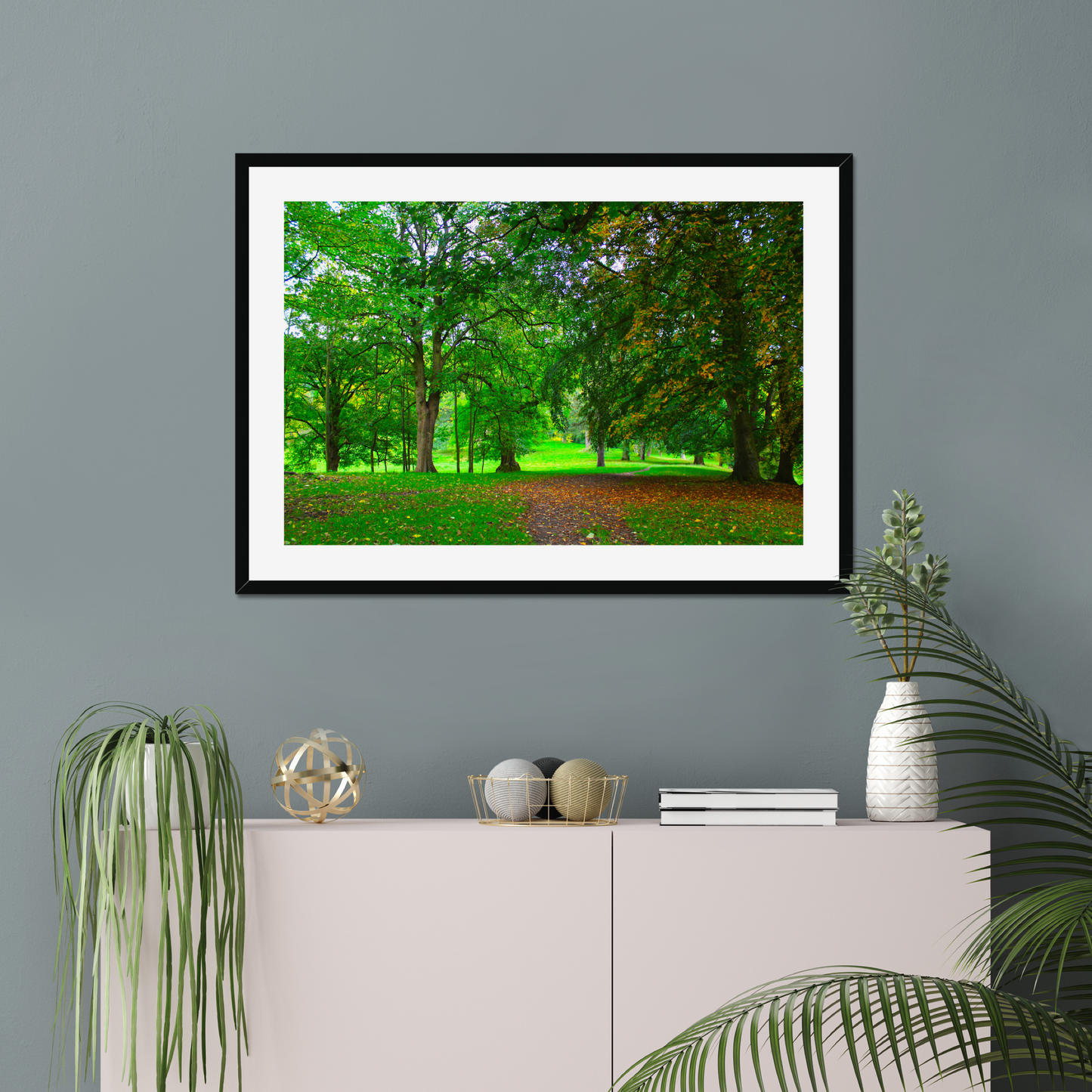 Framed nature print of a green forest scene on a gray wall above a white cabinet with decorative items.