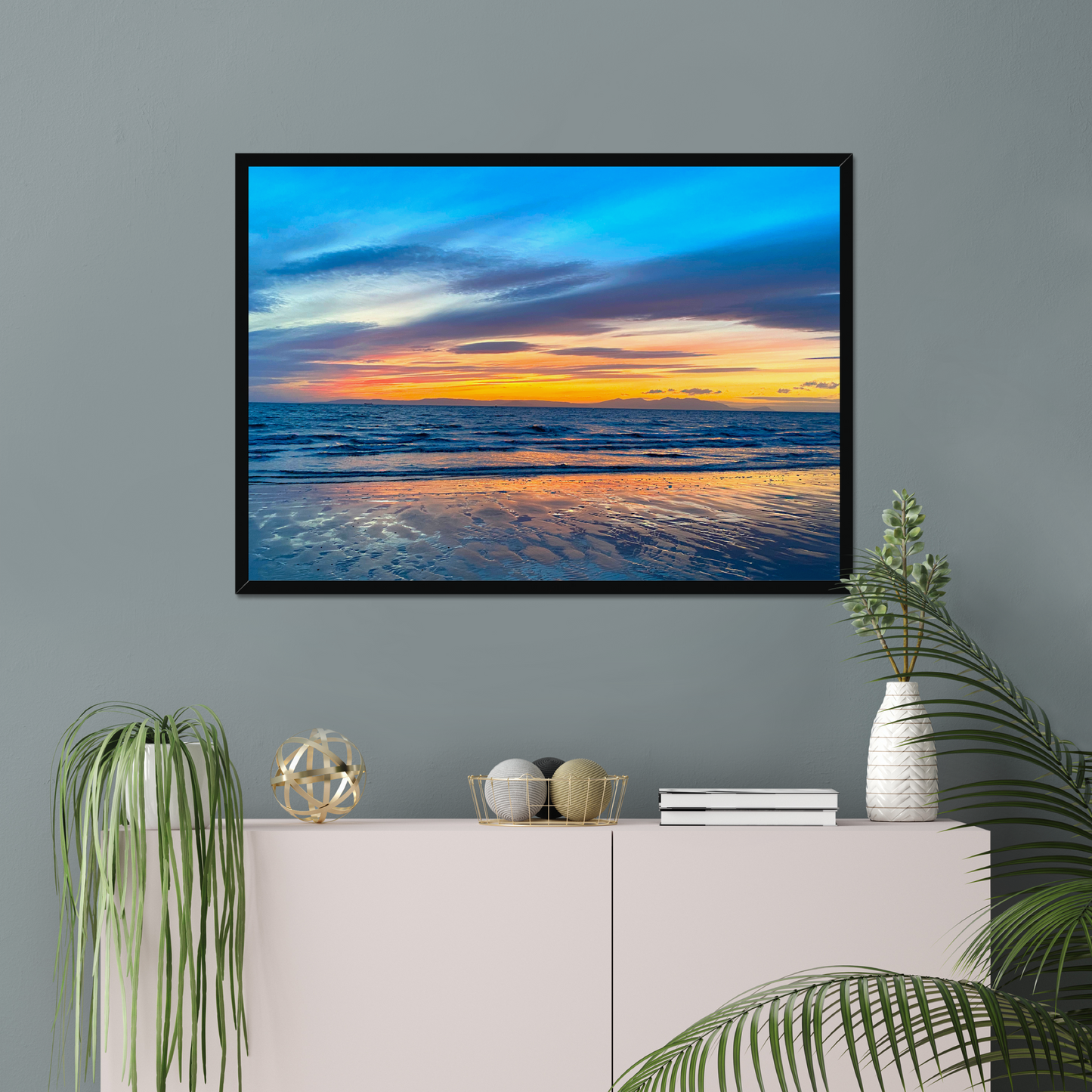 Framed artwork of a sunset over water on a gray wall above a white console table with decorative items.