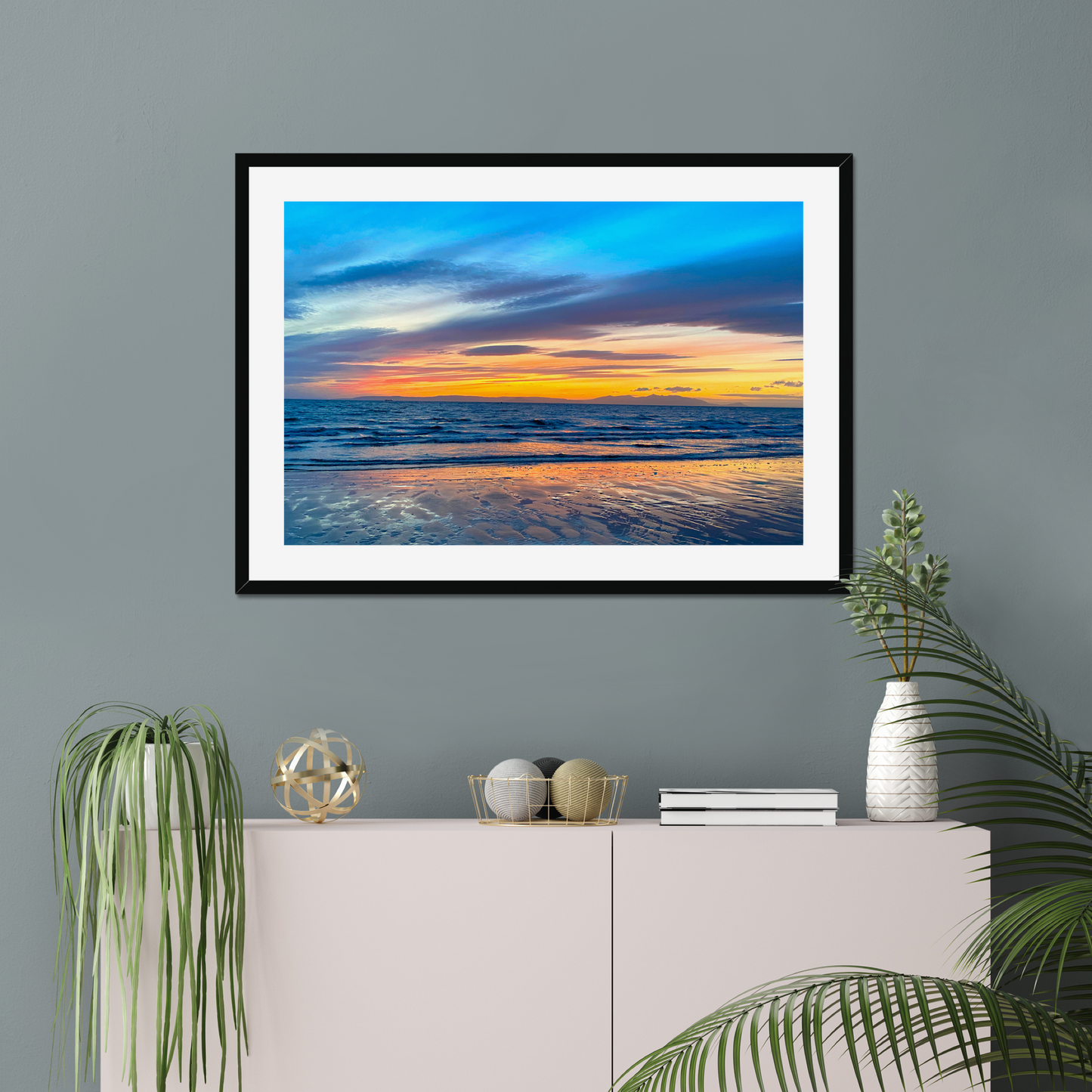 Framed artwork of a sunset over water on a gray wall above a white console table with decorative items.