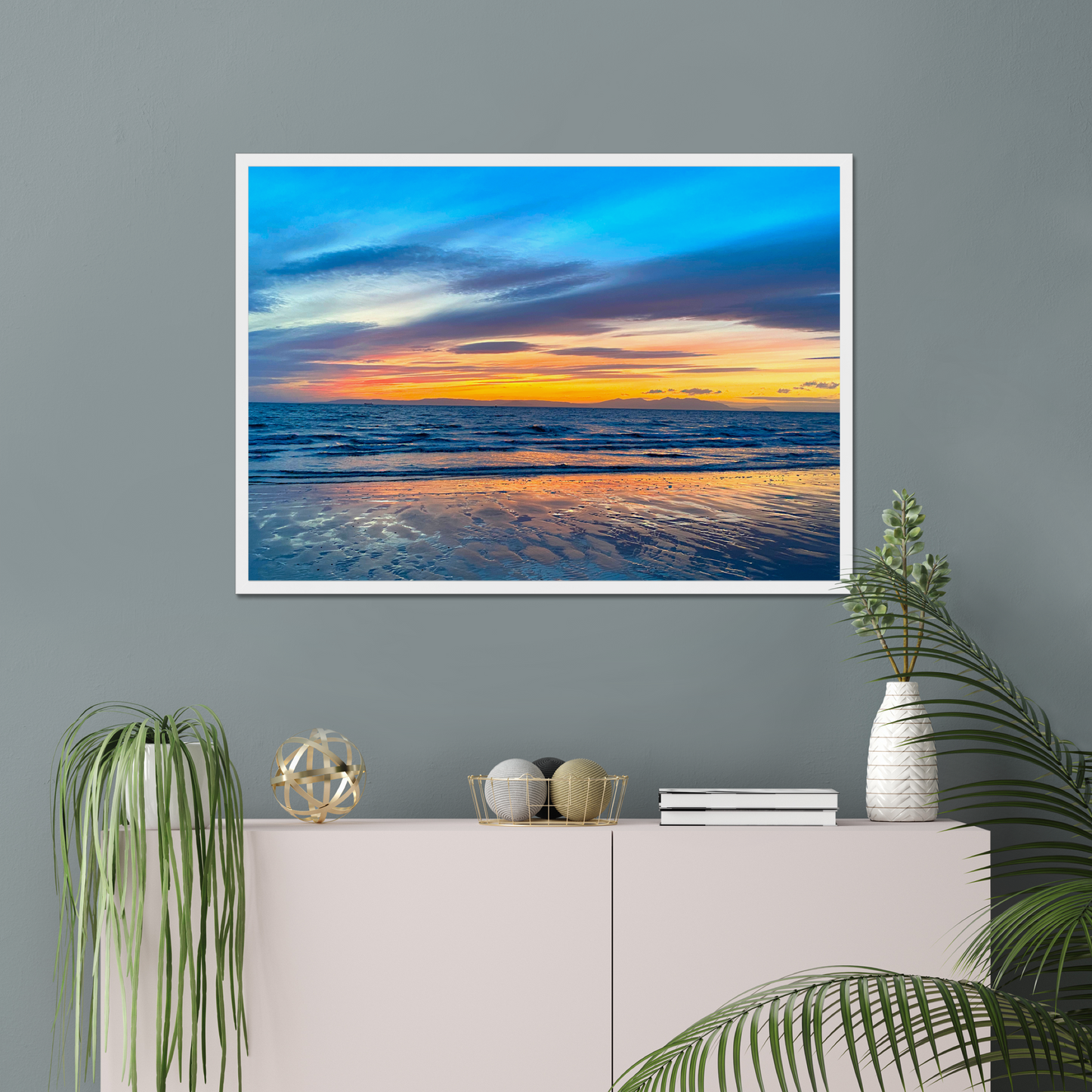 Framed artwork of a sunset over water on a gray wall above a white console table with decorative items.