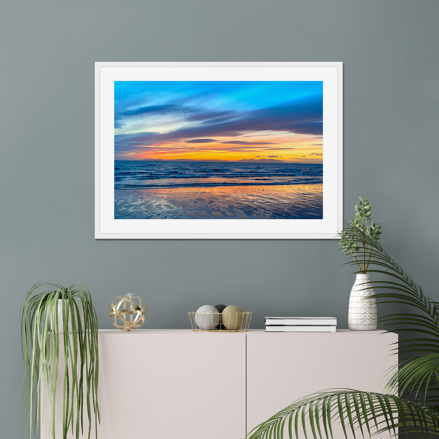 Framed artwork of a sunset over water on a gray wall above a white console table with decorative items.