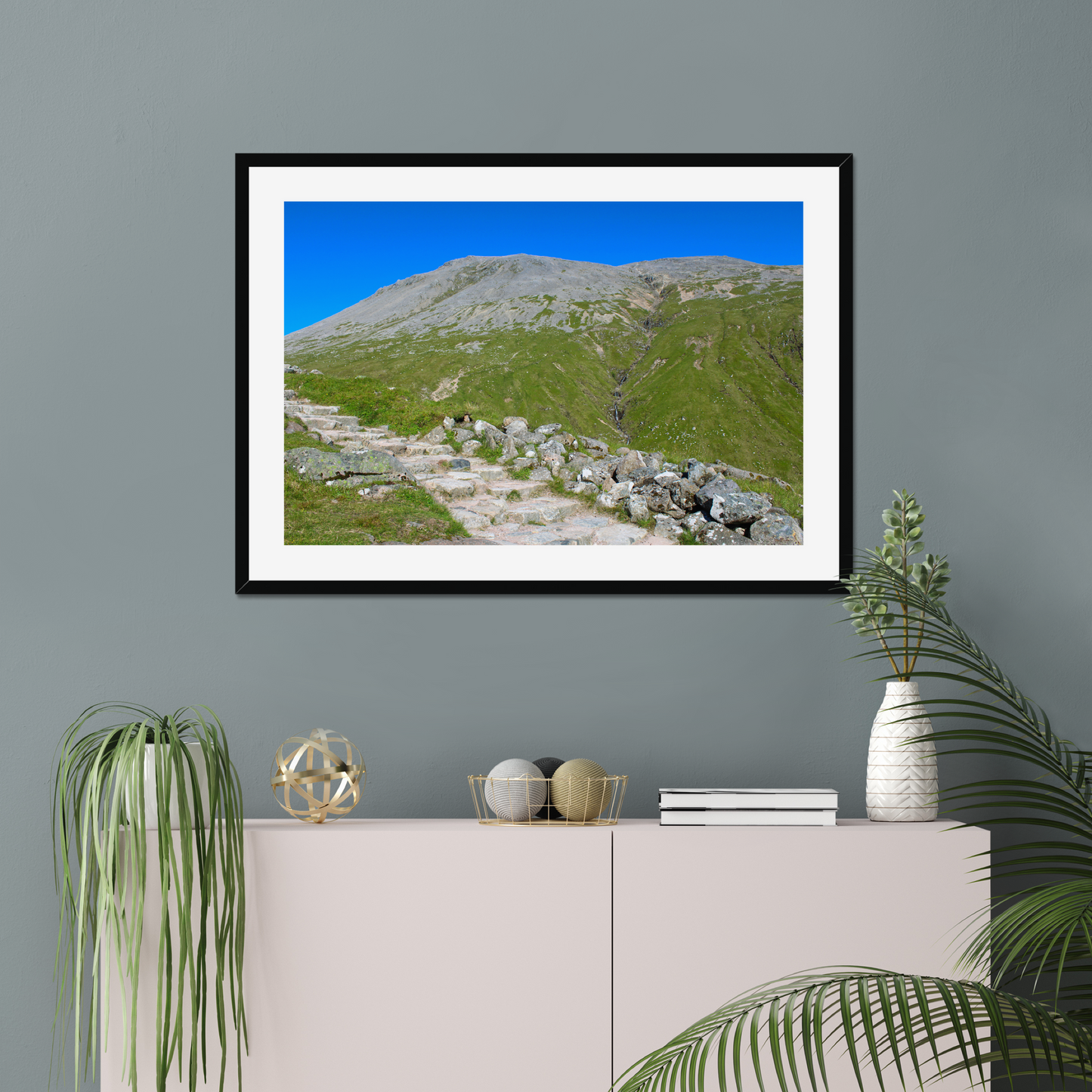 Framed landscape print of a mountainous area with greenery and clear blue sky, displayed on a gray wall above a white cabinet.
