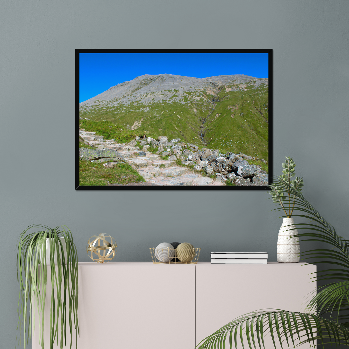 Framed landscape print of a mountainous area with greenery and clear blue sky, displayed on a gray wall above a white cabinet.