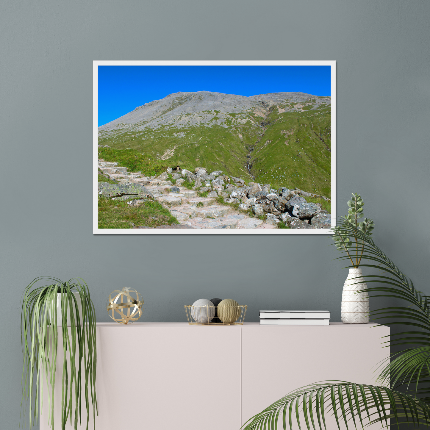Framed landscape print of a mountainous area with greenery and clear blue sky, displayed on a gray wall above a white cabinet.
