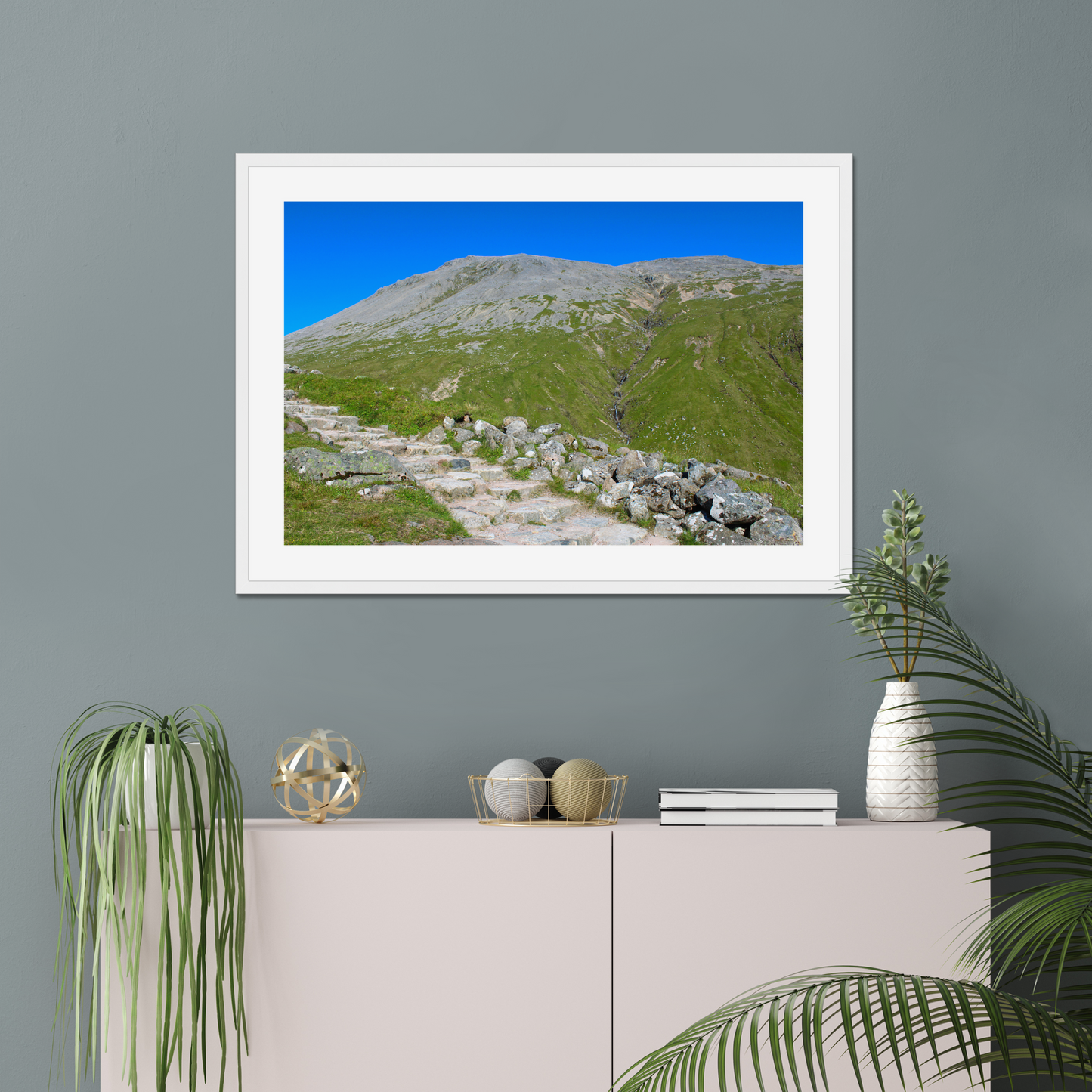 Framed landscape print of a mountainous area with greenery and clear blue sky, displayed on a gray wall above a white cabinet.