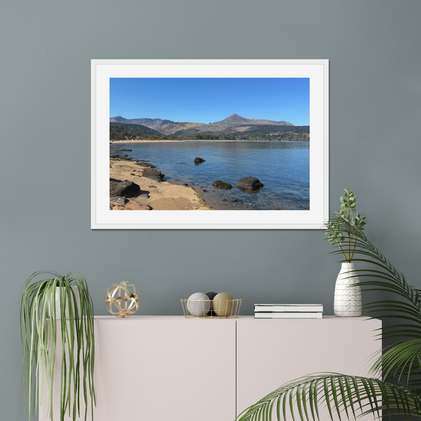 Framed photograph of a coastal scene with mountains in the background on a gray wall above a white cabinet.