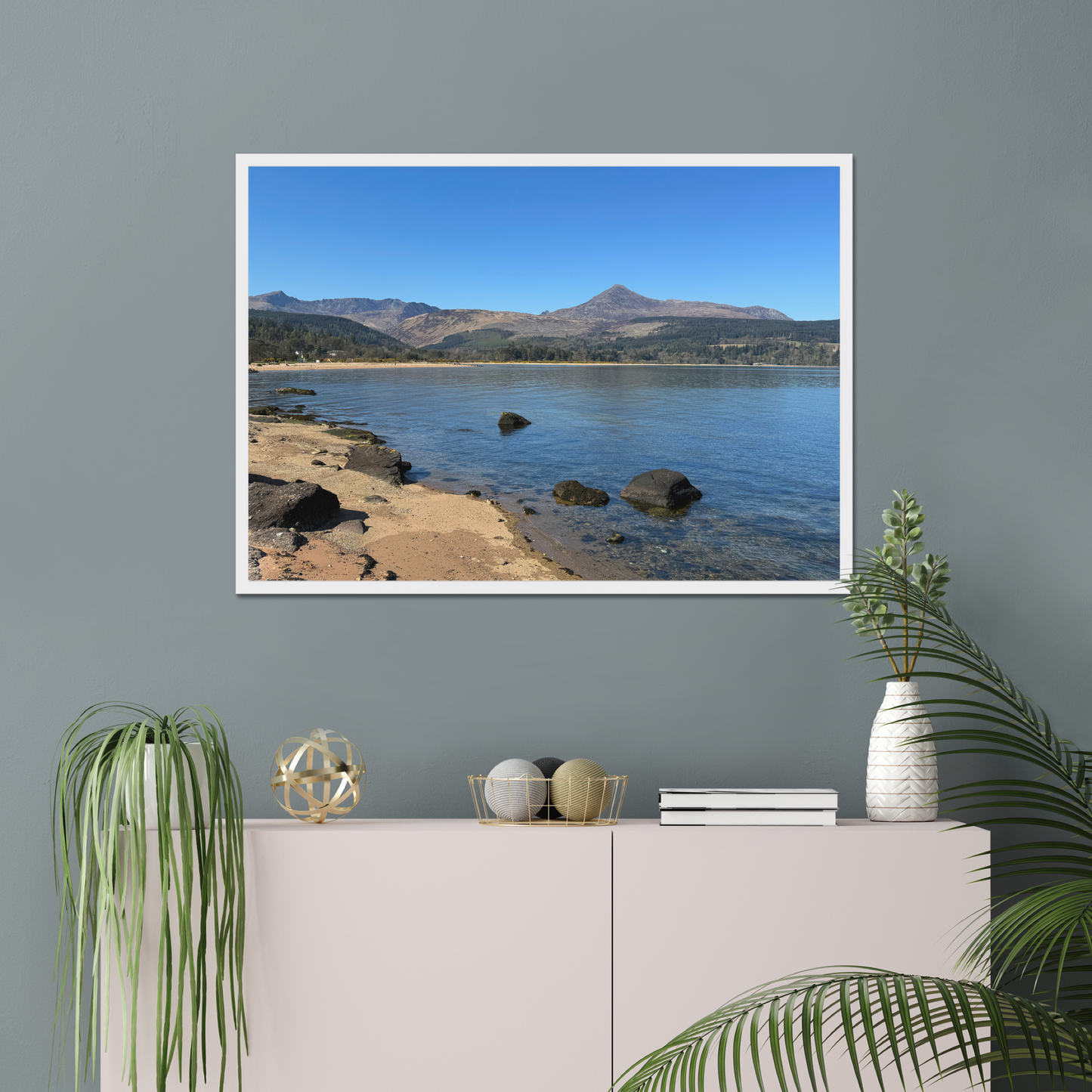 Framed photograph of a coastal scene with mountains in the background on a gray wall above a white cabinet.