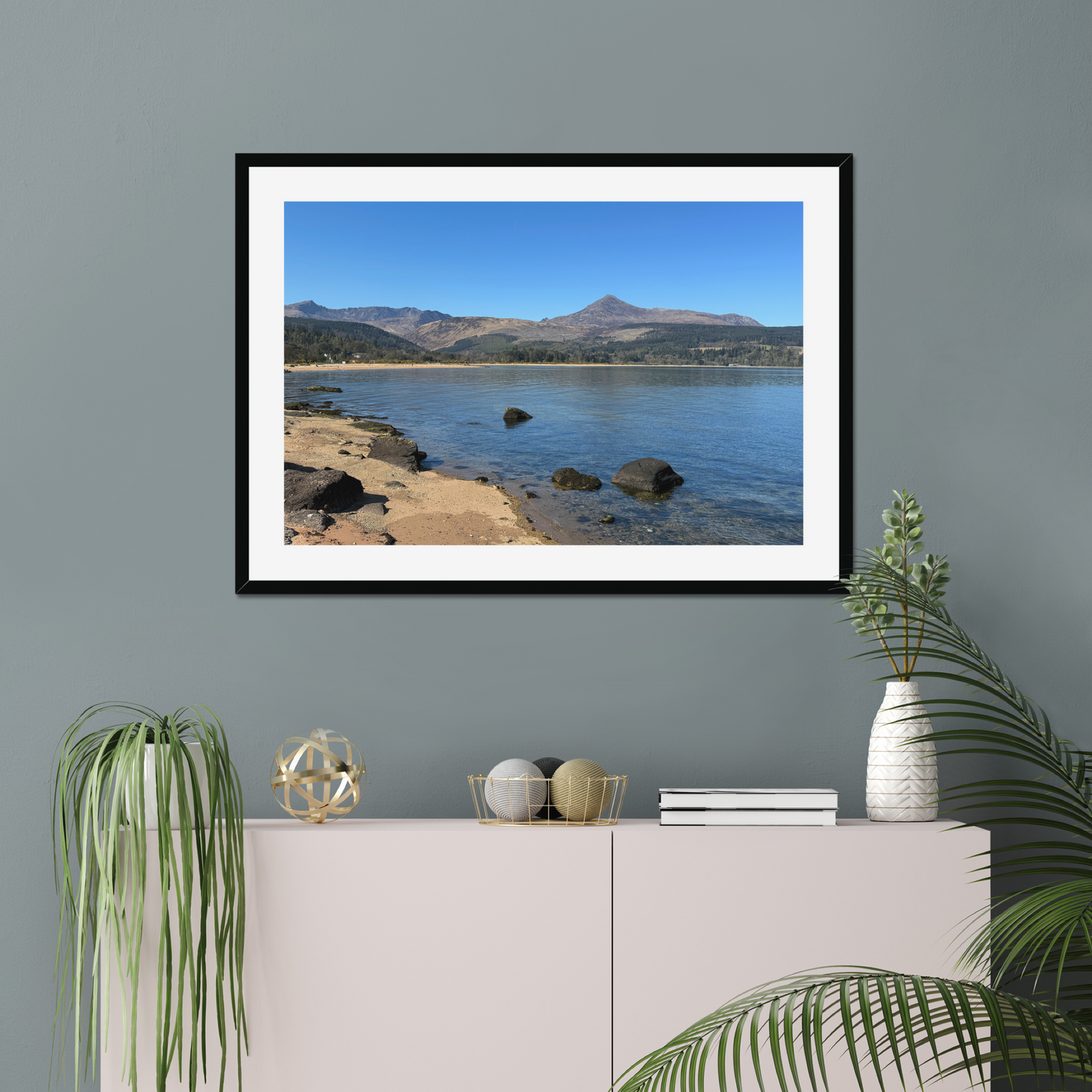 Framed photograph of a coastal scene with mountains in the background on a gray wall above a white cabinet.