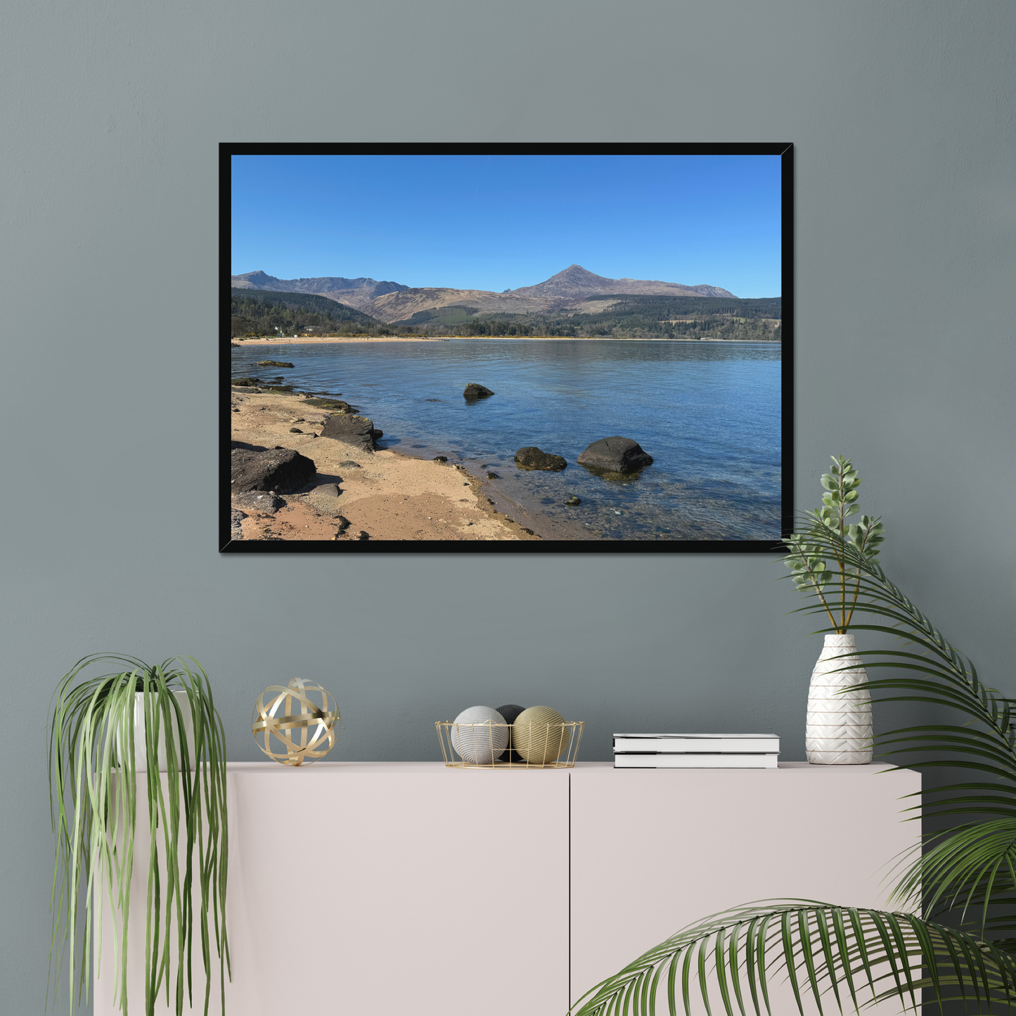 Framed photograph of a coastal scene with mountains in the background on a gray wall above a white cabinet.