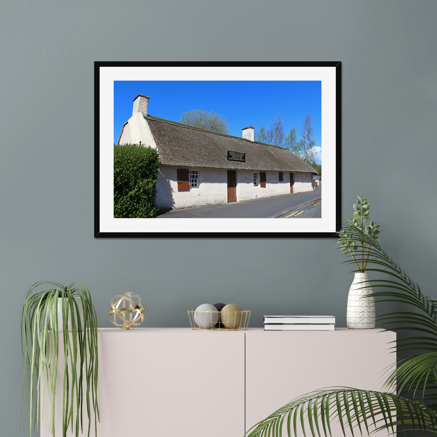 Framed picture of a thatched cottage with a clear blue sky on a gray wall above a white cabinet.