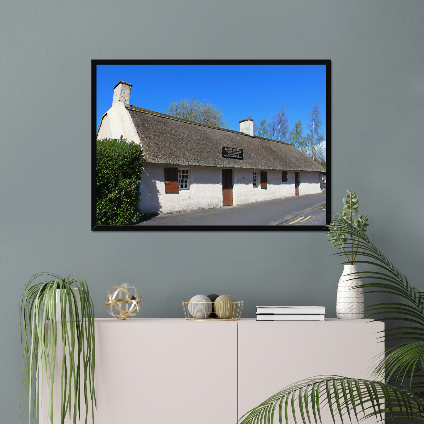 Framed picture of a thatched cottage with a clear blue sky on a gray wall above a white cabinet.