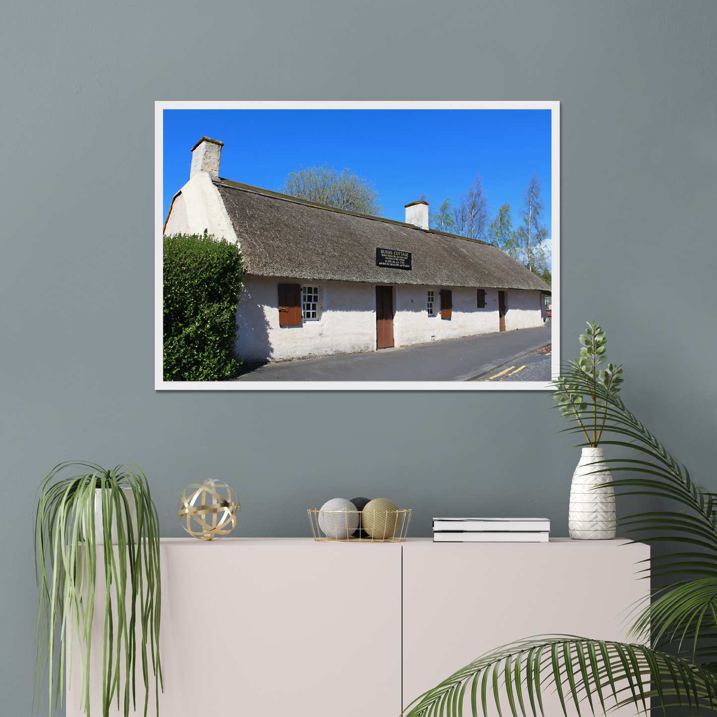 Framed picture of a thatched cottage with a clear blue sky on a gray wall above a white cabinet.