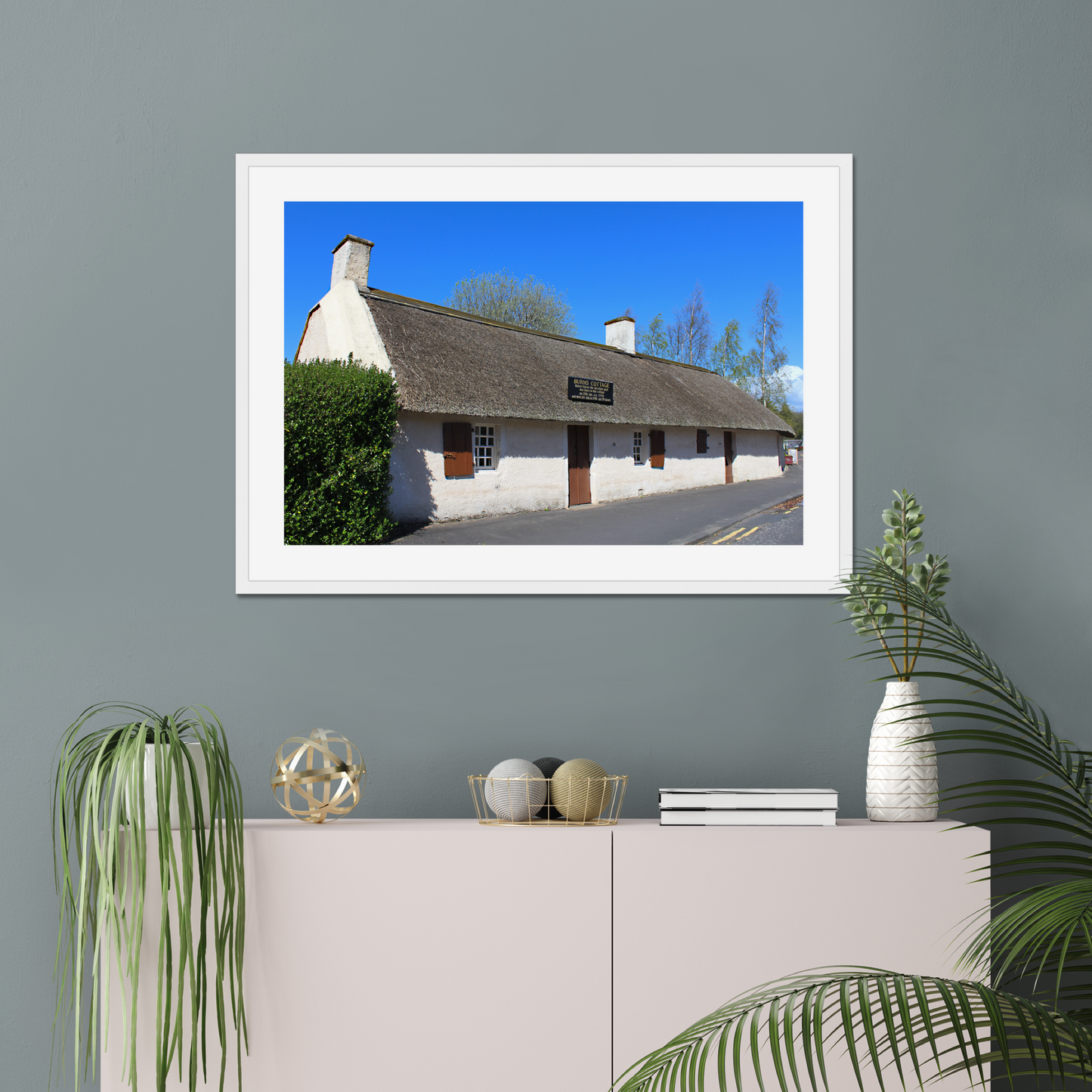 Framed picture of a thatched cottage with a clear blue sky on a gray wall above a white cabinet.