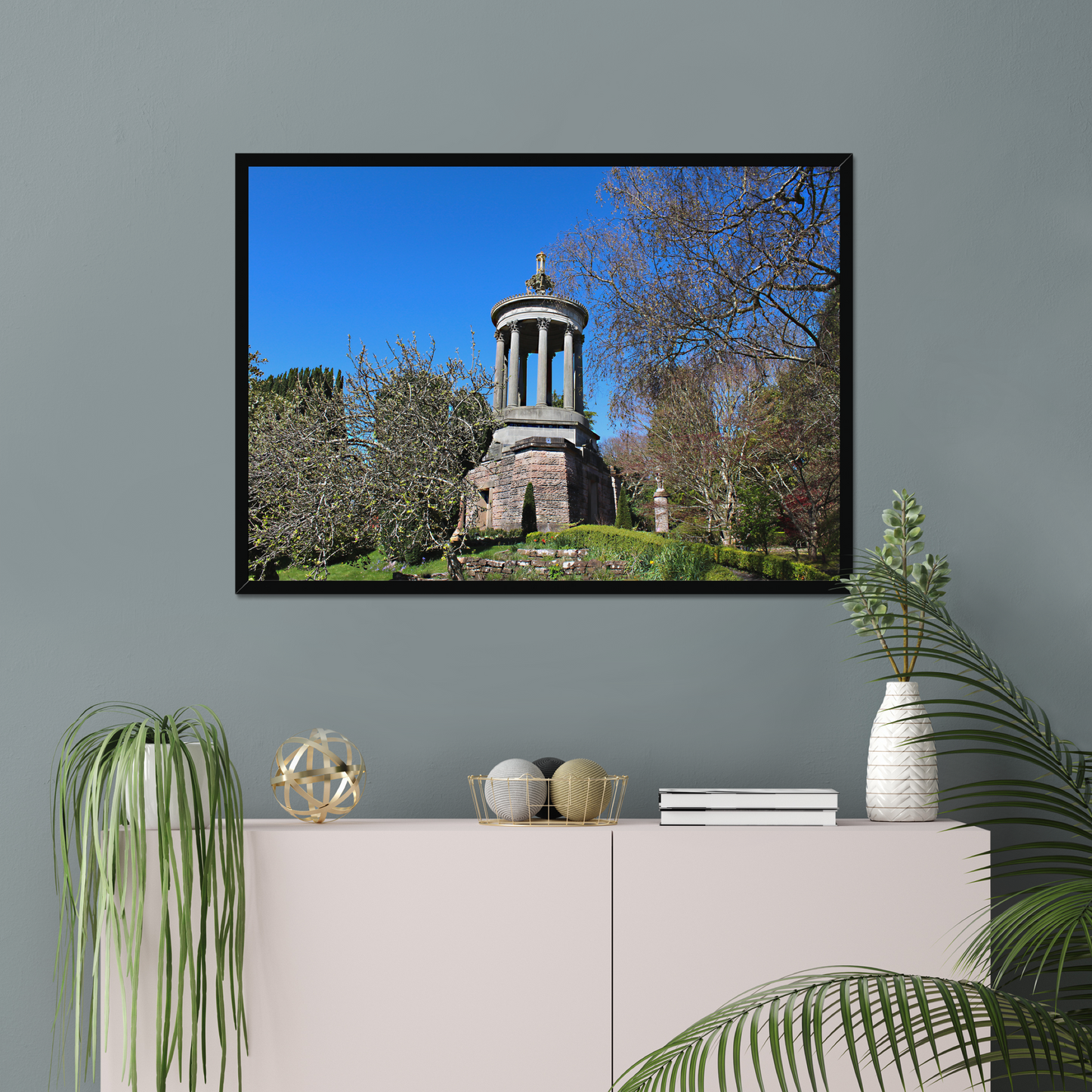 Framed photograph of a historical monument with trees against a blue sky, displayed on a wall above a cabinet with decorative items.