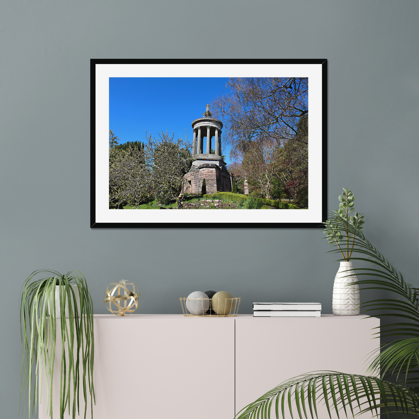Framed photograph of a historical monument with trees against a blue sky, displayed on a wall above a cabinet with decorative items.