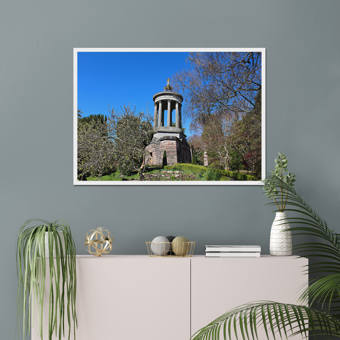 Framed photograph of a historical monument with trees against a blue sky, displayed on a wall above a cabinet with decorative items.