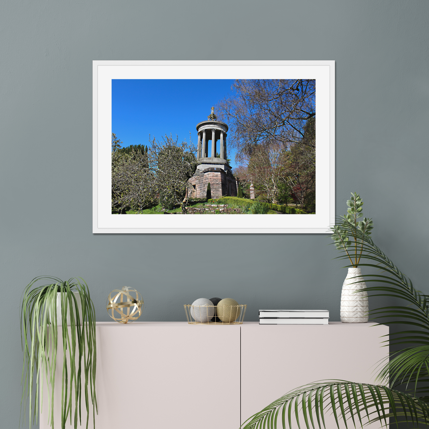 Framed photograph of a historical monument with trees on a gray wall above a white cabinet with decorative items.