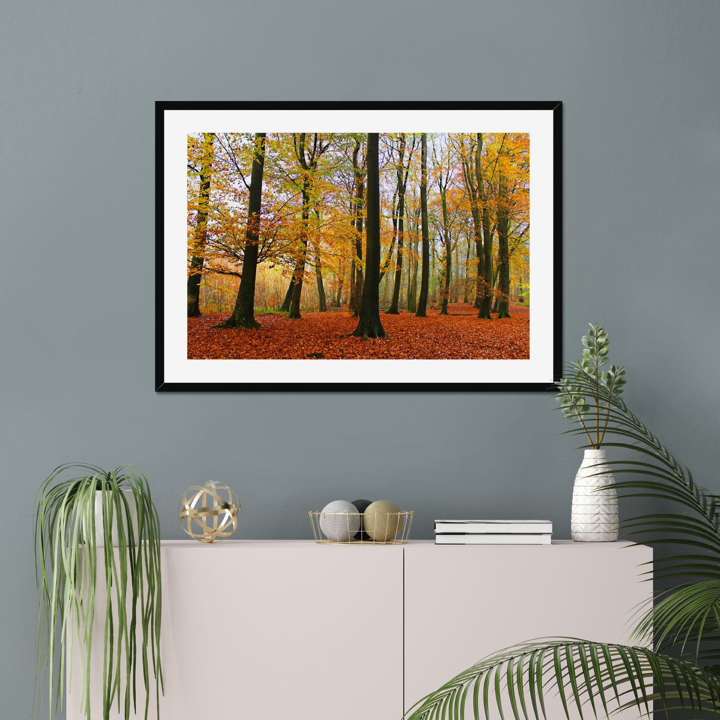 Framed print of a forest scene with autumn colors on a gray wall above a white cabinet.