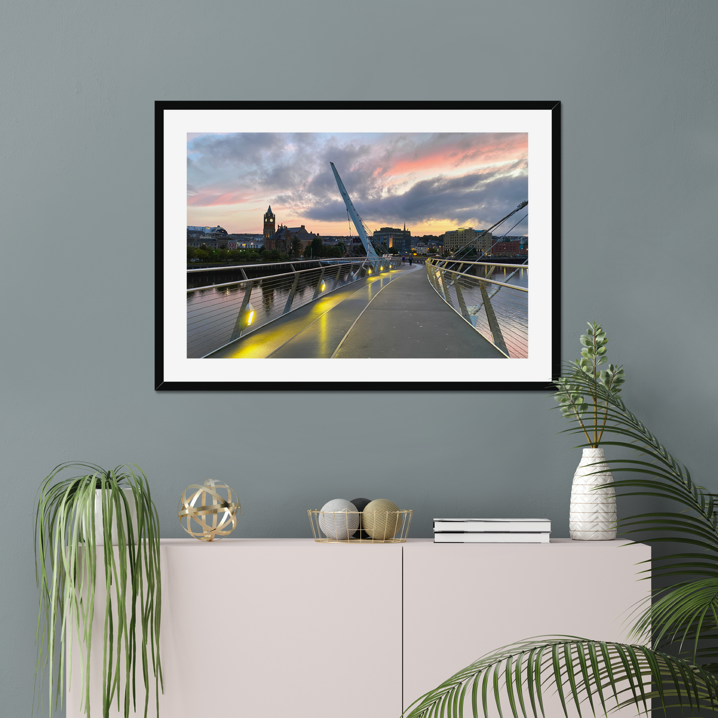 Framed artwork of a bridge at sunset on a gray wall above a white cabinet with decorative items.