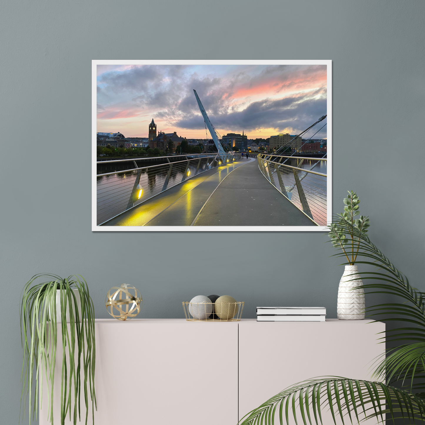 Framed artwork of a cityscape with a bridge against a sunset sky, displayed on a wall above a white cabinet.
