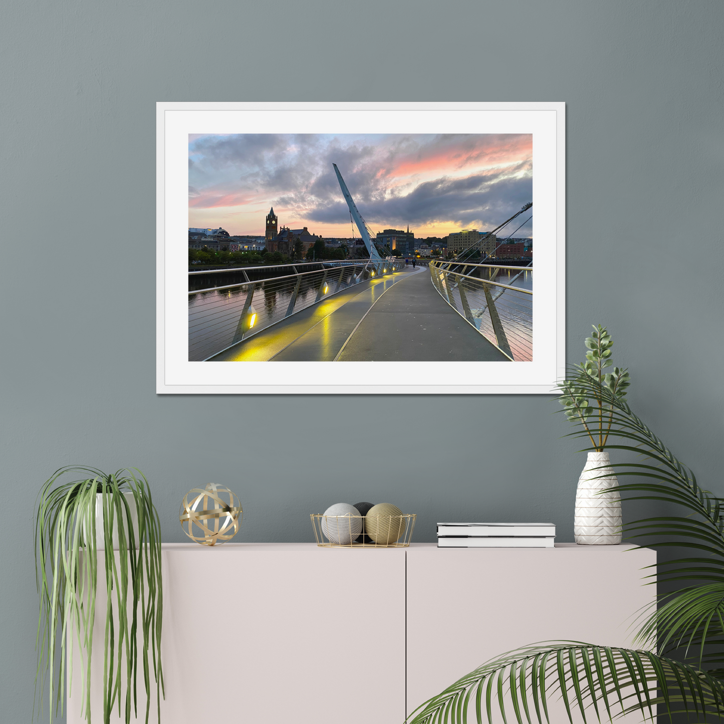 Framed photograph of a bridge with a sunset over a cityscape, displayed on a wall above a white cabinet with decorative items.