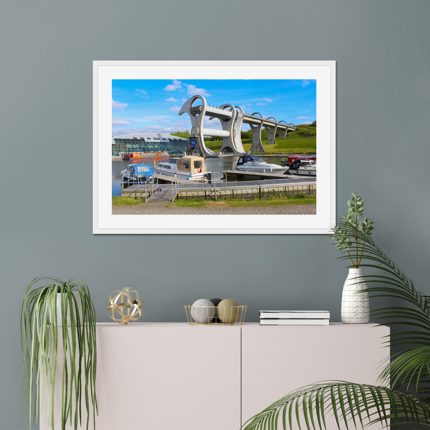 Framed picture of a bridge with boats on a gray wall above a white cabinet with decorative items.