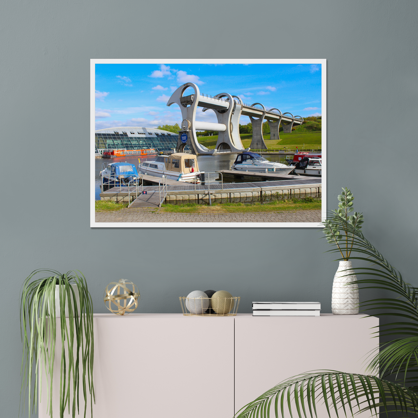 Framed picture of a bridge with boats on a gray wall above a white cabinet with decorative items.