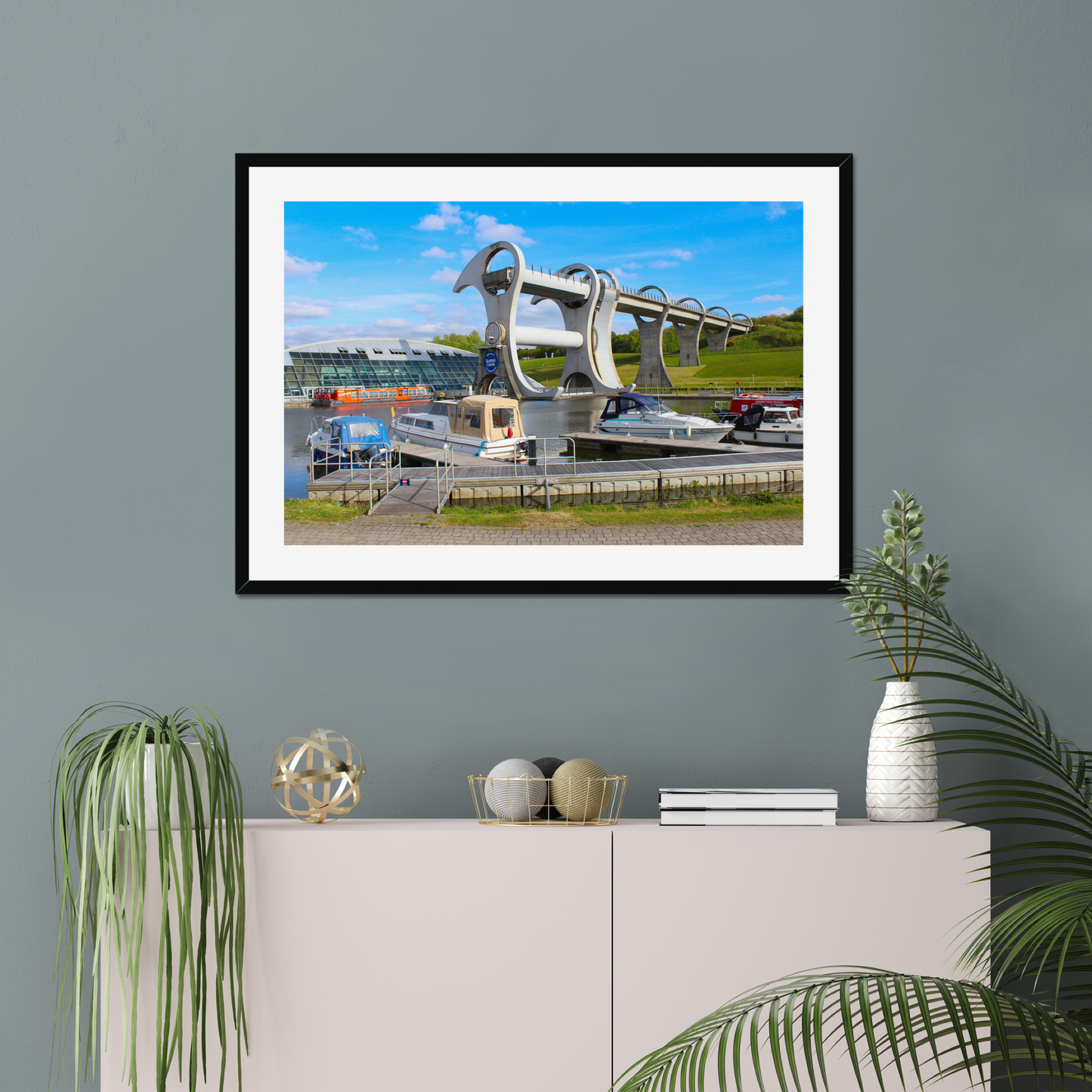 Framed picture of a bridge with boats on a gray wall above a white cabinet with decorative items.