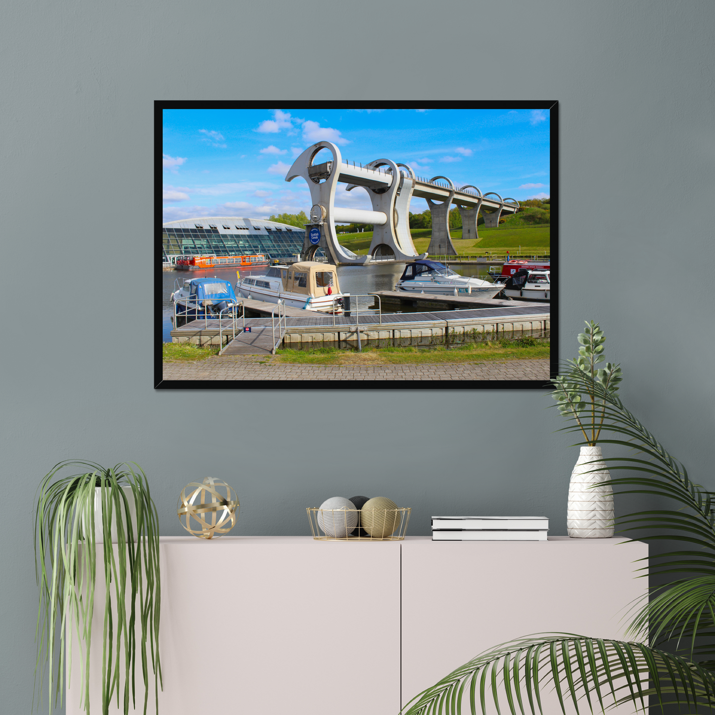 Framed picture of a bridge with boats on a gray wall above a white cabinet with decorative items.