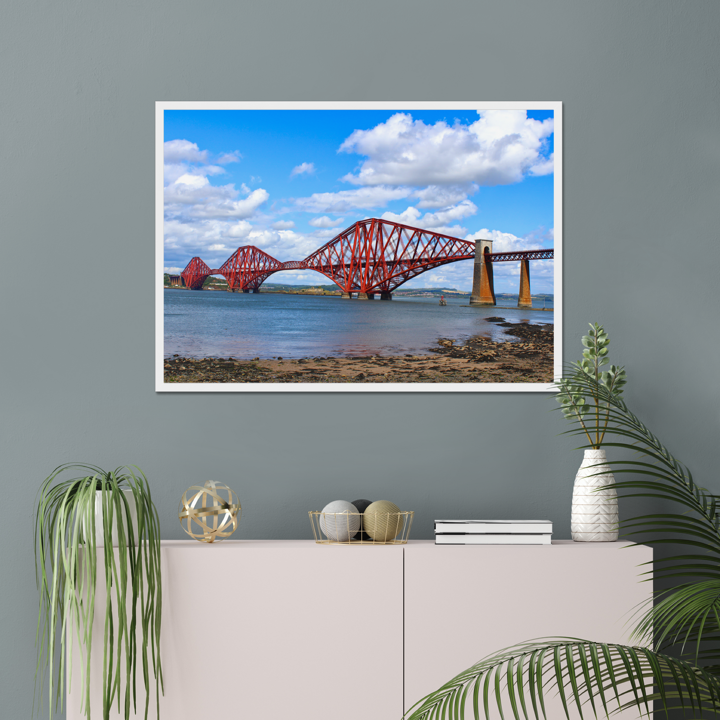 Framed artwork of a coastal scene with a bridge and water on a gray wall above a white cabinet with decorative items.