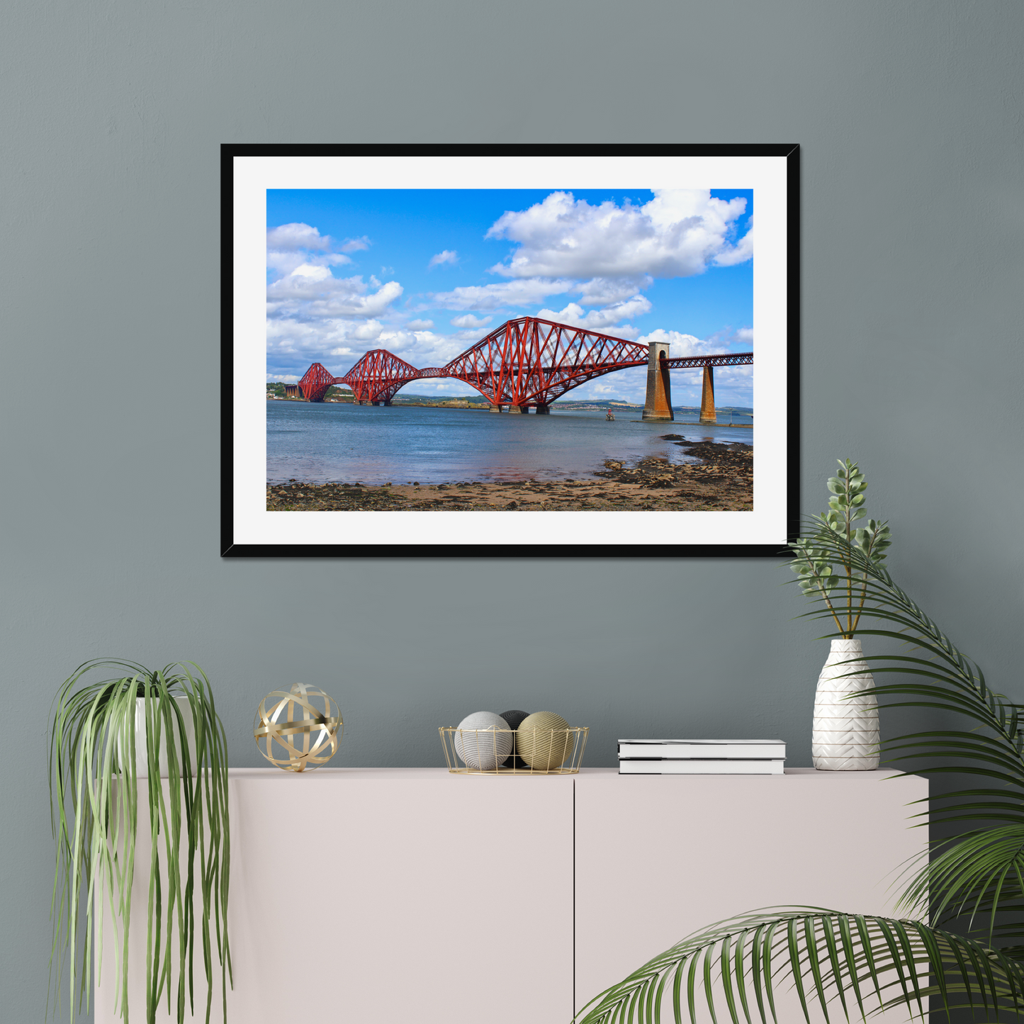 Framed artwork of a coastal scene with a bridge and water on a gray wall above a white cabinet with decorative items.