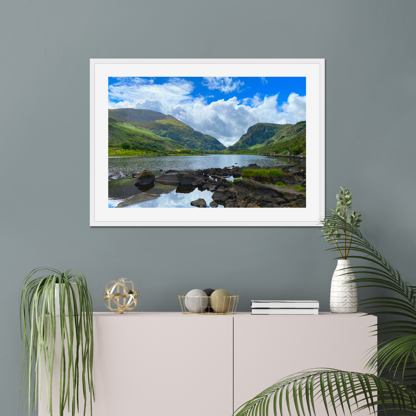 Framed photograph of a lake with mountains in the background on a gray wall with decorative items below.