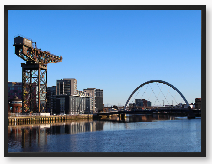 Framed Photo Of A Cityscape with a crane, buildings, and a bridge over water.