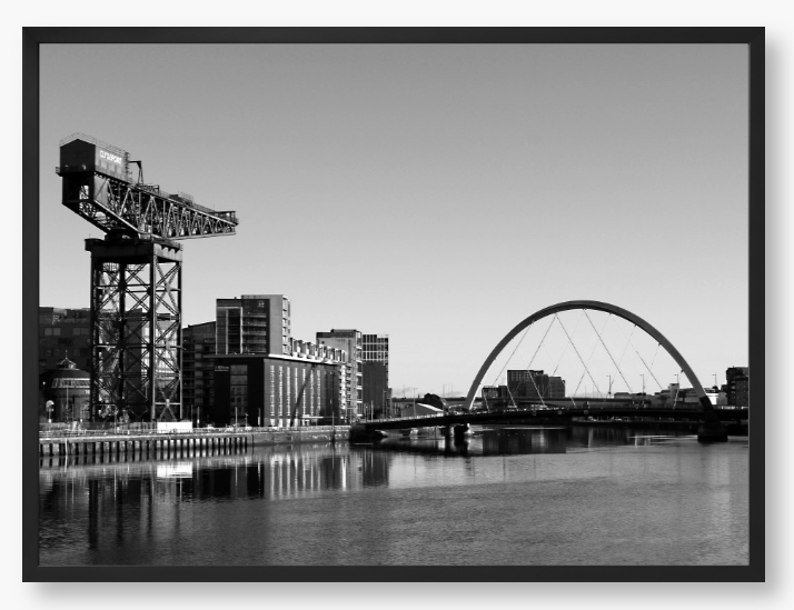 Black and White Framed Photo Of A Cityscape with a crane, buildings, and a bridge over water.