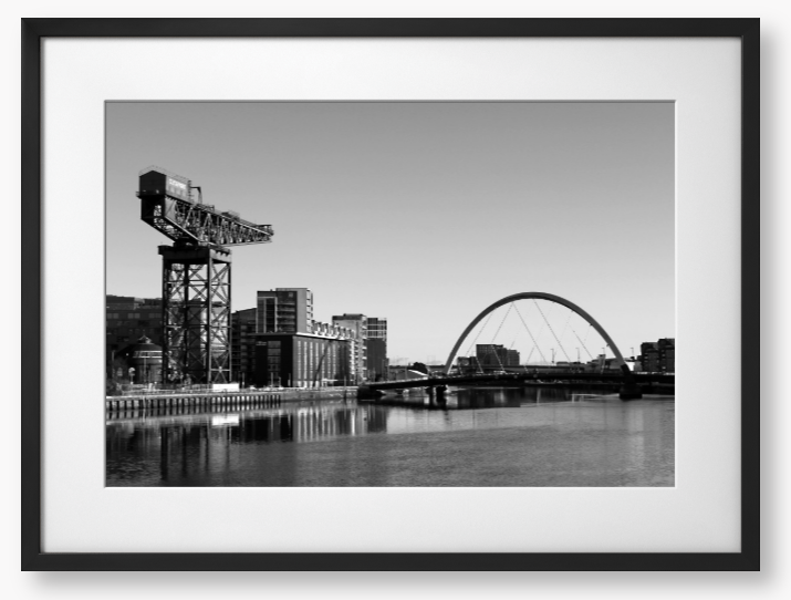 Black and White Framed Photo Of A Cityscape with a crane, buildings, and a bridge over water.