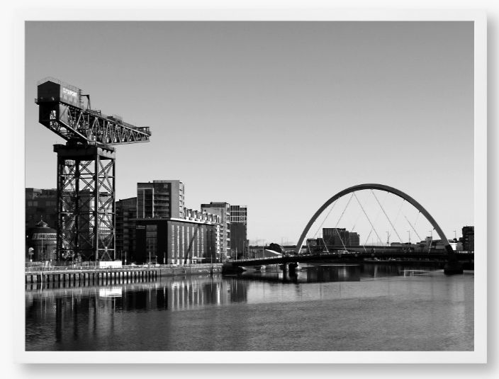 Black and White Framed Photo Of A Cityscape with a crane, buildings, and a bridge over water.