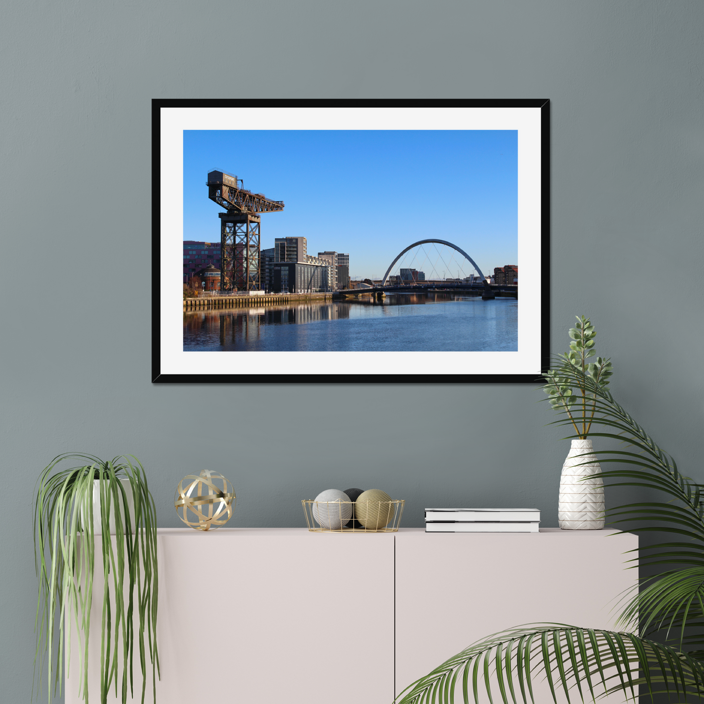 Framed photograph of a cityscape with a crane and a bridge over water hanging on a wall above a white cabinet with decorative items.