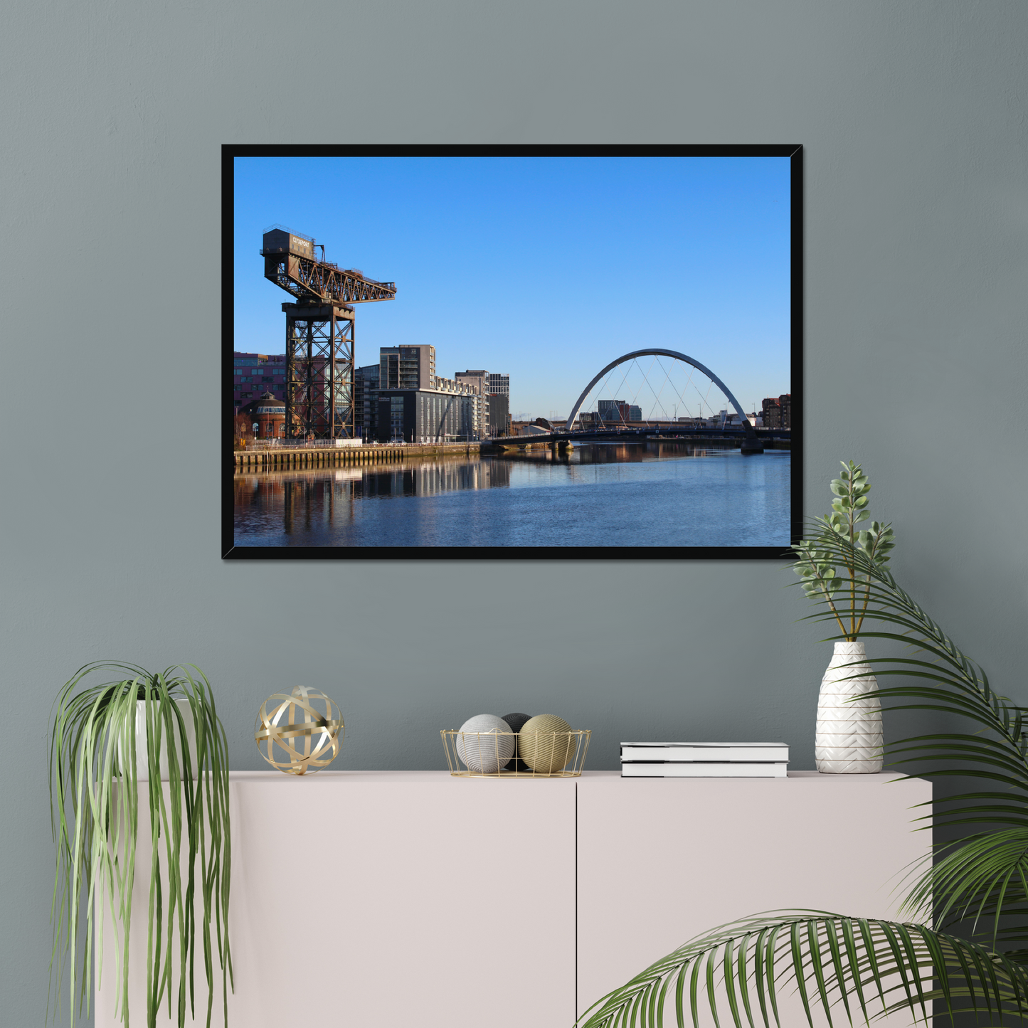 Framed photograph of a cityscape with a crane and a bridge over water hanging on a wall above a white cabinet with decorative items.