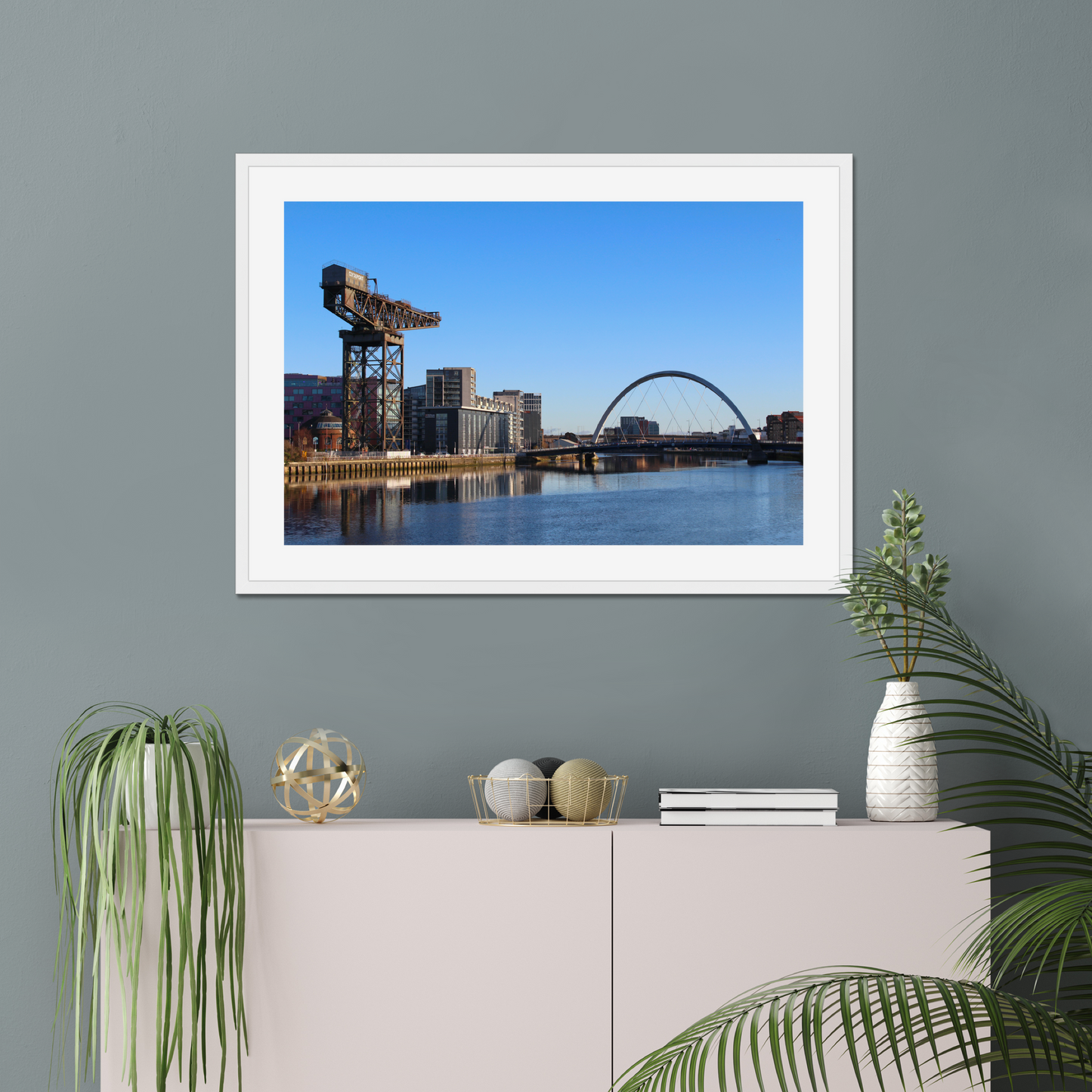 Framed photograph of a cityscape with a crane and a bridge over water hanging on a wall above a white cabinet with decorative items.