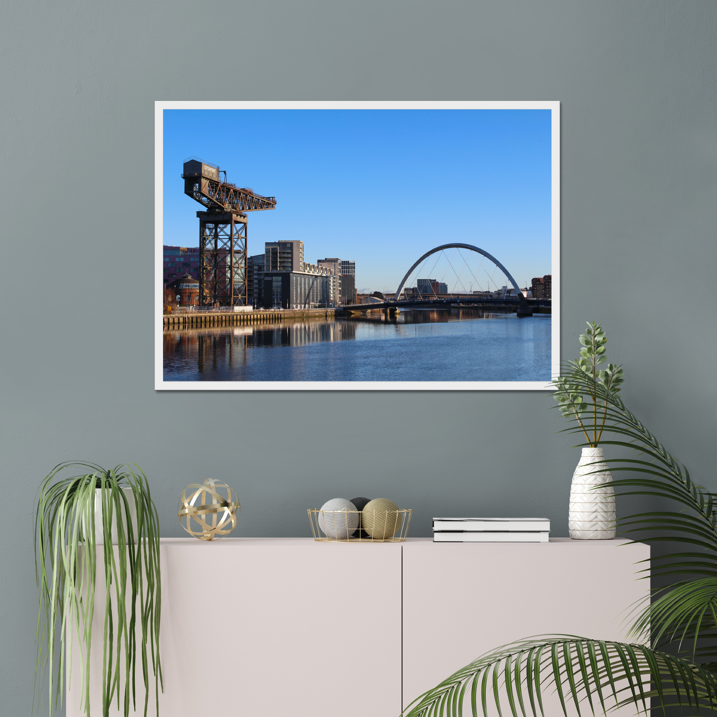 Framed photograph of a cityscape with a crane and a bridge over water hanging on a wall above a white cabinet with decorative items.