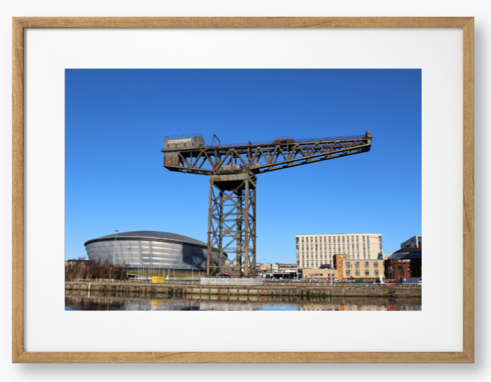Framed Photo of a Industrial crane with modern buildings in the background under a clear blue sky.