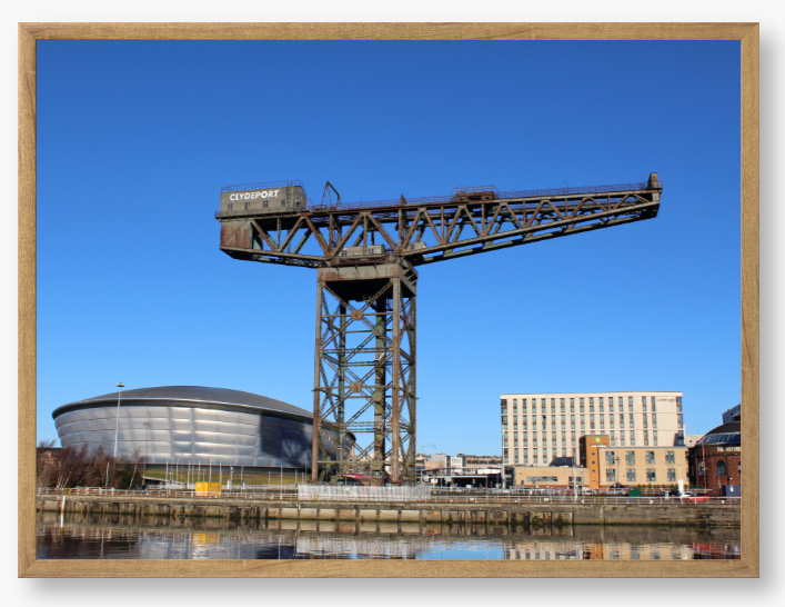 Framed Photo of a Industrial crane with modern buildings in the background under a clear blue sky.