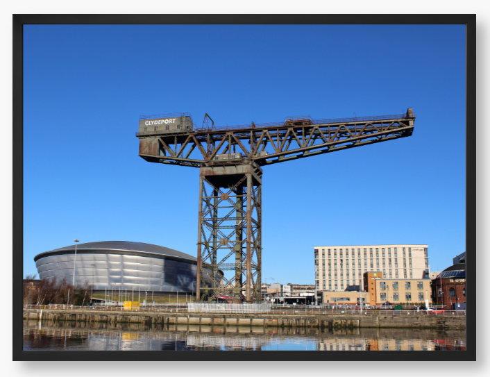 Framed Photo of a Industrial crane with modern buildings in the background under a clear blue sky.