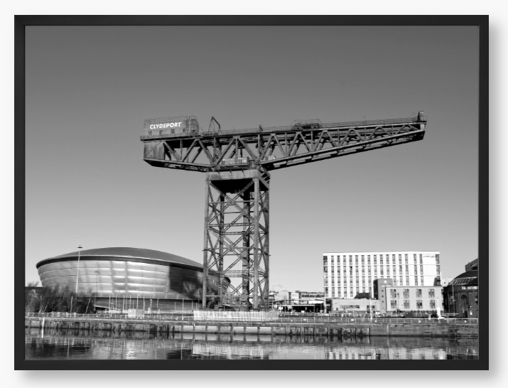 Black and white framed photo of a large crane with modern buildings in the background.