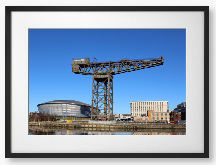 Framed Photo of a Industrial crane with modern buildings in the background under a clear blue sky.